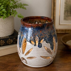 May include: A ceramic candle holder with a unique design. The top is a deep blue and brown, transitioning to white with leaf-shaped cutouts. The interior glows with warm light, creating a cozy ambiance. A decorative piece for home decor.