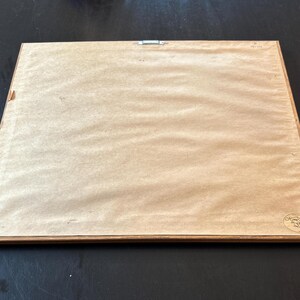 Pu&ograve; includere: Il retro di un'opera d'arte incorniciata, con un supporto di carta marrone chiaro e una cornice in legno. Un gancio di metallo &egrave; fissato nella parte superiore. La cornice &egrave; rettangolare e sembra vintage.