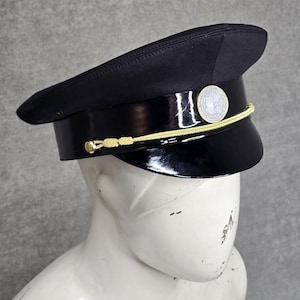 May include: A black peaked cap featuring a shiny black visor and a black band. A thin gold cord runs across the front, secured by small gold buttons on each side. A circular silver emblem is centered on the front band above the visor.