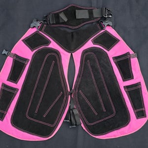 May include: Pink and black chaps with pockets and adjustable straps. The chaps feature black suede-like panels with pink stitching, designed to protect the legs. The item is set against a black background.
