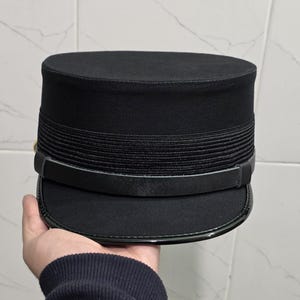 Op de afbeelding: Een zwarte hoed in militaire stijl met een platte bovenkant en een korte, gebogen rand. De hoed heeft een geribbelde band rond de kroon en een glanzende zwarte band boven de rand. De hoed wordt in een hand gehouden.