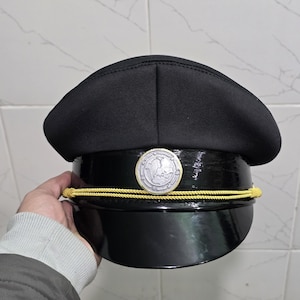 Może przedstawiać: Czarny kapelusz z błyszczącym, zaokrąglonym rondem i matową górą. Srebrny emblemat jest wyśrodkowany z przodu, nad złotym sznurkiem. Kapelusz jest trzymany przez osobę.