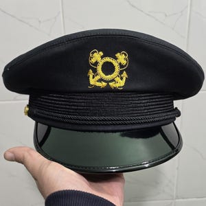 May include: Black military-style hat with a shiny, dark green visor. The hat features a gold emblem and gold buttons. The hat is held in a hand.