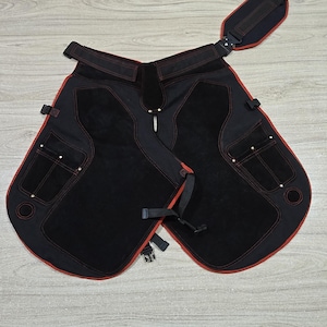 May include: Black leather work apron with red stitching and trim. Features include pockets, adjustable straps, and a padded neck strap. The apron is designed for protection and utility in a workshop or similar environment.