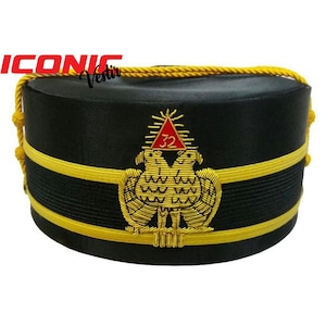 Puede incluir: Sombrero masónico negro con ribete dorado y un emblema bordado en oro que representa dos águilas y un triángulo rojo con el número 32.
