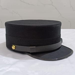 Pode incluir: Um boné preto de topo plano com uma coroa estruturada e detalhes canelados horizontais na base. Uma tira de aspeto de couro castanho escuro envolve a parte inferior, fixada por um botão metálico de tom dourado polido no lado esquerdo.