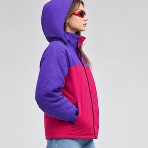 May include: A color-blocked hooded jacket featuring a vibrant purple upper section and a bright pink lower section. The jacket includes a high collar, a front zipper closure, long sleeves with adjustable cuffs, and a hood. Red sunglasses are worn.