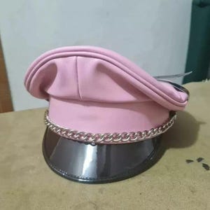 May include: A pink, faux leather hat with a silver chain detail around the base. The hat has a shiny, black visor and a structured crown. The hat is a fashion accessory.