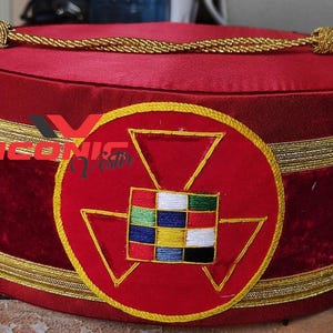 Puede incluir: Sombrero masónico de terciopelo rojo con ribete dorado y un emblema bordado en rojo y dorado que presenta un triángulo y un cuadrado con cuadrados de colores en su interior.