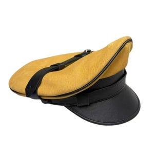 May include: A mustard yellow hat with a black brim and strap. The hat has a rounded top and a structured brim. The black strap goes across the top of the hat. The hat is made of a suede-like material.