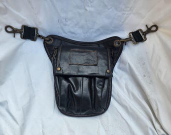Puede incluir: Riñonera de cuero negro con múltiples compartimentos y cierre de solapa. La bolsa presenta un parche rectangular y está adornada con herrajes color latón y una correa negra ajustable con cierres metálicos.
