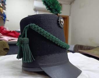 May include: A black shako hat featuring a dark green feather plume, a silver-toned star-shaped emblem, and a braided dark green cord with matching tassels. The hat includes a black visor and a black fabric band around the base of the crown.