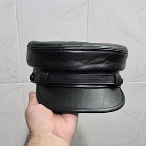 May include: A black leather hat with a flat top and a short, curved brim. The hat has a band around the crown with a buckle detail. The hat is held in a hand against a tiled background.