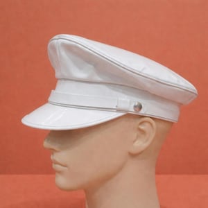 May include: A white, glossy, military-style hat with a visor and a band around the crown. The hat has a silver button detail on the side. The hat is set against an orange background.