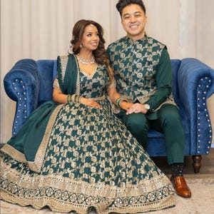 以下が含まれることがあります： お揃いのエメラルドグリーンのフォーマルウェアを着たカップル。女性はゴールドのアクセントが施された詳細なスカートとトップスを着用し、男性はグリーンのシャツとパンツの上にマッチするベストを着用しています。彼らは青いベルベットのソファに座っています。