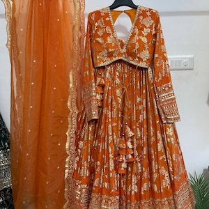 Könnte beinhalten: Ein orangefarbenes und goldenes indisches Brautkleid mit einem langärmeligen Oberteil, einem Rock mit Blumenmuster und einem durchsichtigen Dupatta. Das Kleid ist mit goldenen Stickereien und Pailletten verziert.