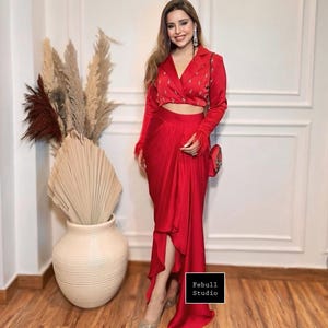 May include: A striking red two-piece outfit comprising a cropped blazer with feather trim and a matching draped skirt. The blazer has decorative embroidery. A small red purse and silver heels complete the ensemble. The outfit is from Febull Studio.