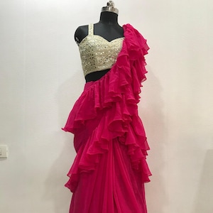 以下が含まれることがあります： ゴールドの装飾が施されたクロップトップが付いたピンクのフリルサリ。サリは伝統的なスタイルで、複数のフリルを重ねてドレープされています。