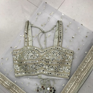 以下が含まれることがあります： 金色の縁取りとスパンコールが付いた白いネット生地。白いミラーワークとスパンコールのクロップトップが生地の上に置かれています。