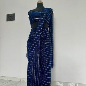May include: A navy blue saree with silver horizontal stripes is displayed on a mannequin. The saree is draped to showcase the design, with a matching blouse and long sleeve. The fabric appears to have a slight sheen.