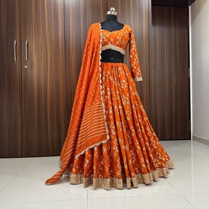 Könnte beinhalten: Ein orangefarbenes und golden besticktes Lehenga mit passendem Dupatta. Das Lehenga hat einen Blumendruck und einen goldenen Rand. Das Dupatta hat ein gestreiftes Muster und einen gewellten Rand.