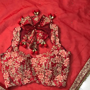 以下が含まれることがあります： 金糸とスパンコールで刺繍された赤いブライダルブラウス。ブラウスは丸首でノースリーブのデザイン。ブラウスは、金色の縁取りのある赤いシアーデュパッタと組み合わせています。