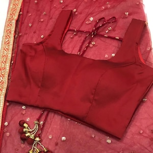 以下が含まれることがあります： 金と赤の刺繍が縁取りされた赤いネットのサリー。サリーには、丸首の赤いブラウスが付属しています。