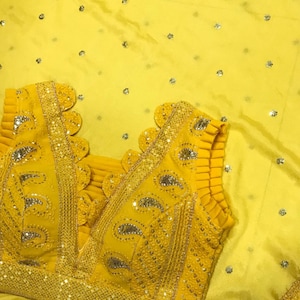 以下が含まれることがあります： 鮮やかな黄色のブラウスで、複雑なスパンコールと刺繍のディテールが施されています。 ブラウスは深いVネックとスカラップエッジが特徴です。 生地は微妙な光沢があり、デザインには小さく散らばった装飾が含まれています。