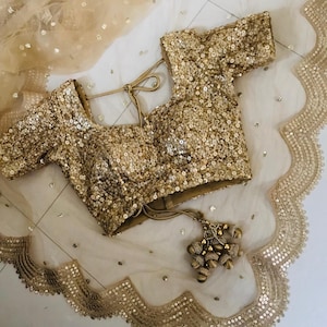 以下が含まれることがあります： 丸首と半袖のゴールドのビーズのブラウス。ブラウスは後ろにリボン留めがあり、小さなゴールドのビーズで飾られています。