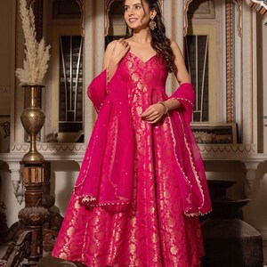 May include: A vibrant fuchsia-colored dress with gold floral patterns. The dress features a V-neckline, a flowing skirt, and matching sheer scarf. The outfit is paired with matching pants. The dress is suitable for special occasions.