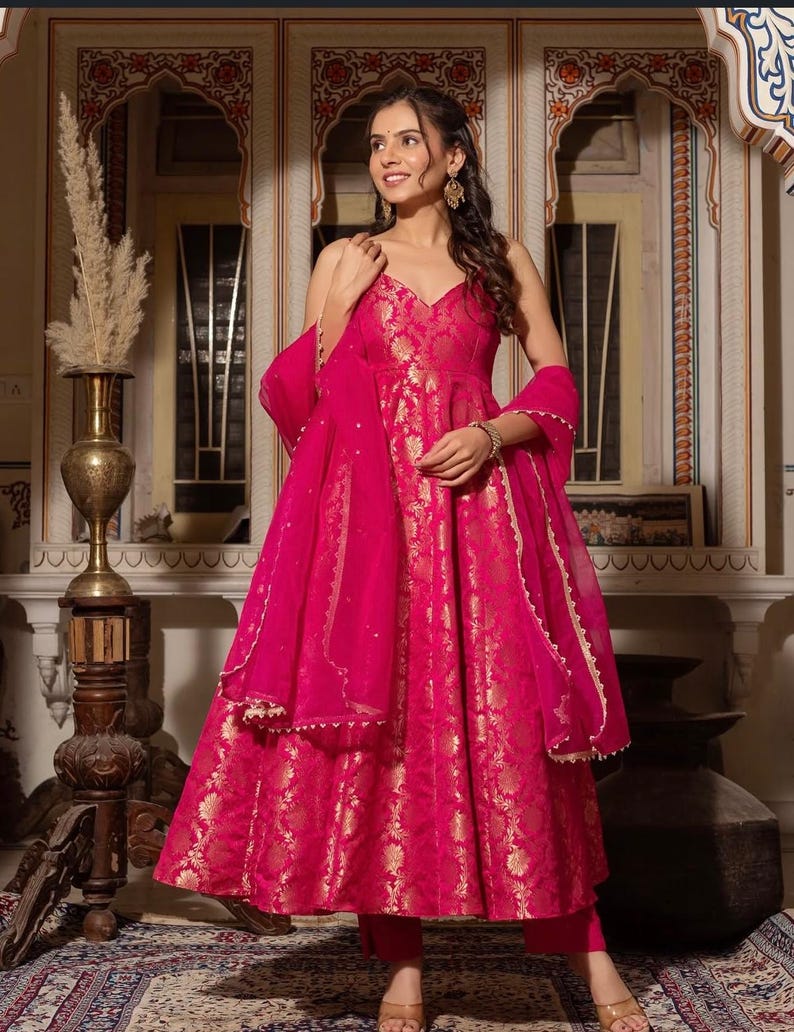 May include: A vibrant fuchsia-colored dress with gold floral patterns. The dress features a V-neckline, a flowing skirt, and matching sheer scarf. The outfit is paired with matching pants. The dress is suitable for special occasions.