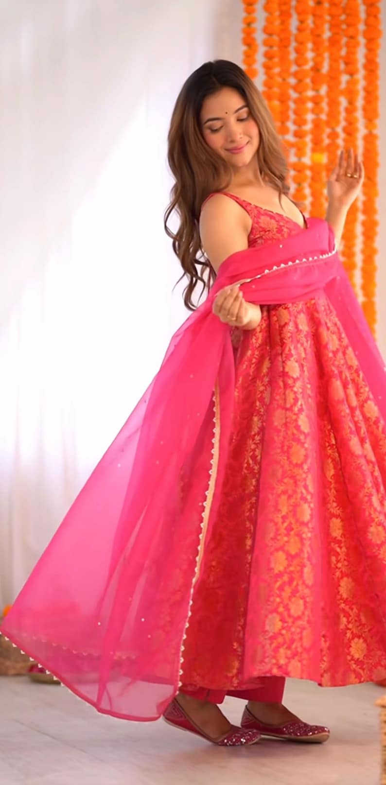 May include: A vibrant pink and gold floral print dress with a matching sheer scarf. The dress features a V-neckline and a flowing skirt. The outfit is paired with matching pink shoes, creating a cohesive and elegant look.