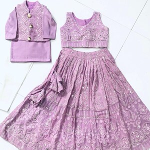 以下が含まれることがあります： 若い女の子のためのライラック色の衣装。袖なしの刺繍入りトップ、フリルのある裾の長いスカート、それに合わせた刺繍入りジャケット。衣装は銀色のスパンコールと刺繍で飾られています。