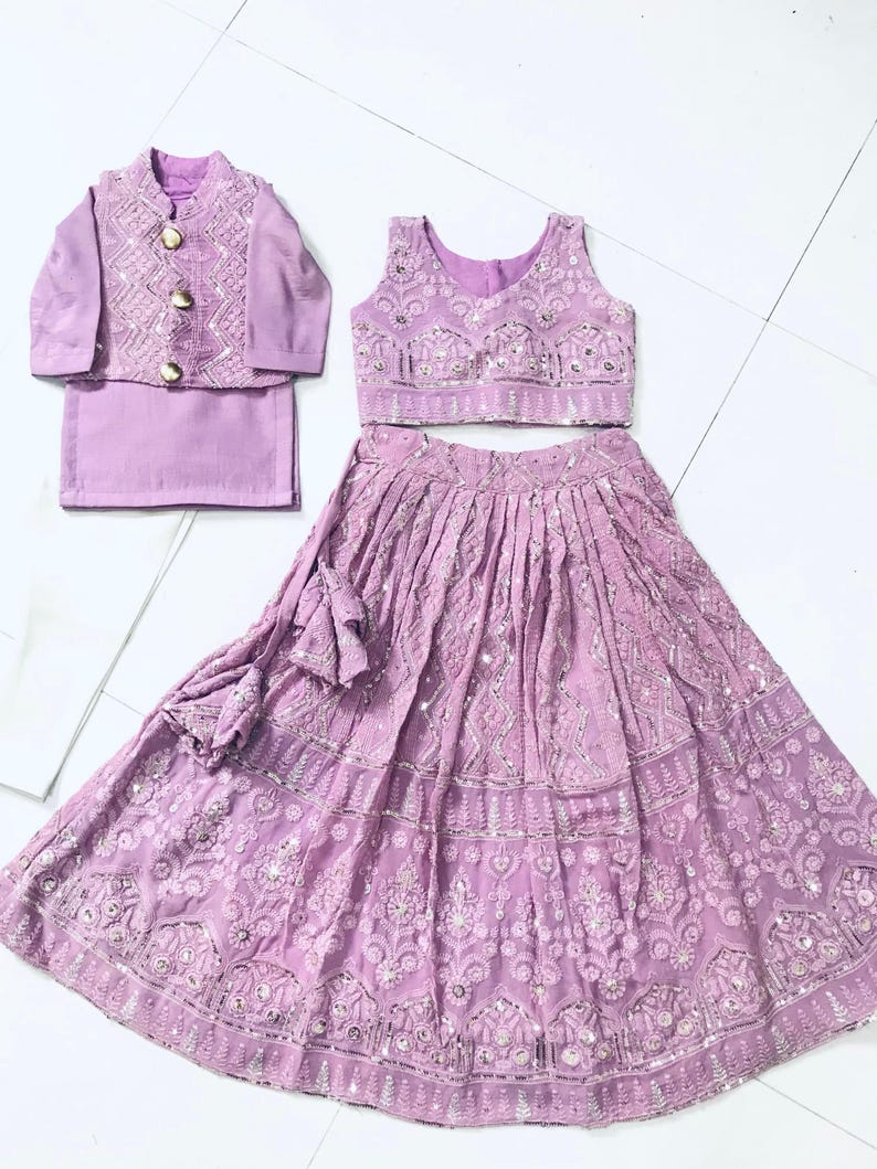 以下が含まれることがあります： 若い女の子のためのライラック色の衣装。袖なしの刺繍入りトップ、フリルのある裾の長いスカート、それに合わせた刺繍入りジャケット。衣装は銀色のスパンコールと刺繍で飾られています。