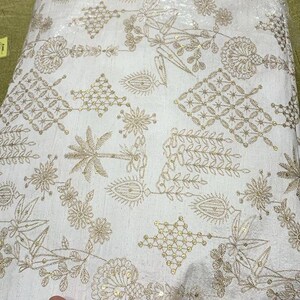 May include: White fabric with intricate gold embroidery. The design features floral and geometric patterns, with small sequins adding sparkle. The fabric is folded, showing the detailed texture and design. Ideal for apparel or decorative projects.