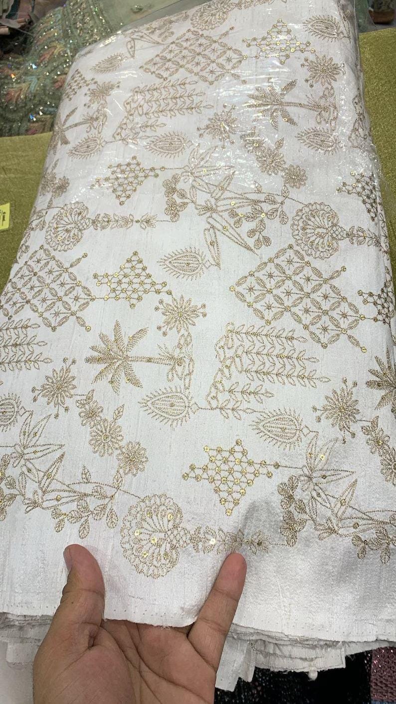 May include: White fabric with intricate gold embroidery. The design features floral and geometric patterns, with small sequins adding sparkle. The fabric is folded, showing the detailed texture and design. Ideal for apparel or decorative projects.