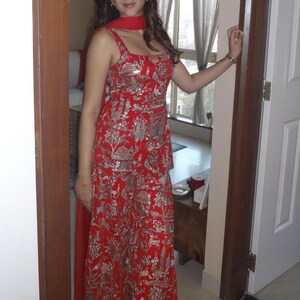 May include: A red sleeveless dress with a detailed silver pattern. The dress has a square neckline and a matching red scarf. The design includes images of buildings, trees, and animals. The dress is worn by a person standing in a doorway.