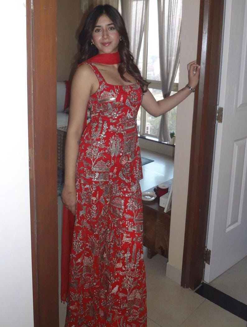 May include: A red sleeveless dress with a detailed silver pattern. The dress has a square neckline and a matching red scarf. The design includes images of buildings, trees, and animals. The dress is worn by a person standing in a doorway.