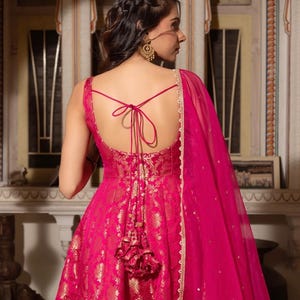 May include: A vibrant fuchsia-colored gown with gold floral brocade detailing. The dress features a low back with tie-up straps and a matching tassel. A sheer fuchsia shawl with gold trim drapes over the shoulder, creating an elegant silhouette.
