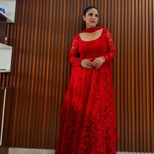May include: A full-length red lace gown with long sleeves and a matching scarf. The dress features a square neckline and a tiered skirt. The background is a wooden wall.