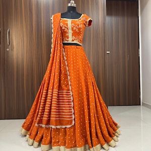 Könnte beinhalten: Ein langer orangefarbener Rock mit goldenen Punkten und einem goldenen Stickrand. Der Rock wird mit einem passenden orangefarbenen Oberteil mit goldener Stickerei und einem passenden orangefarbenen Dupatta mit goldenen Streifen kombiniert.