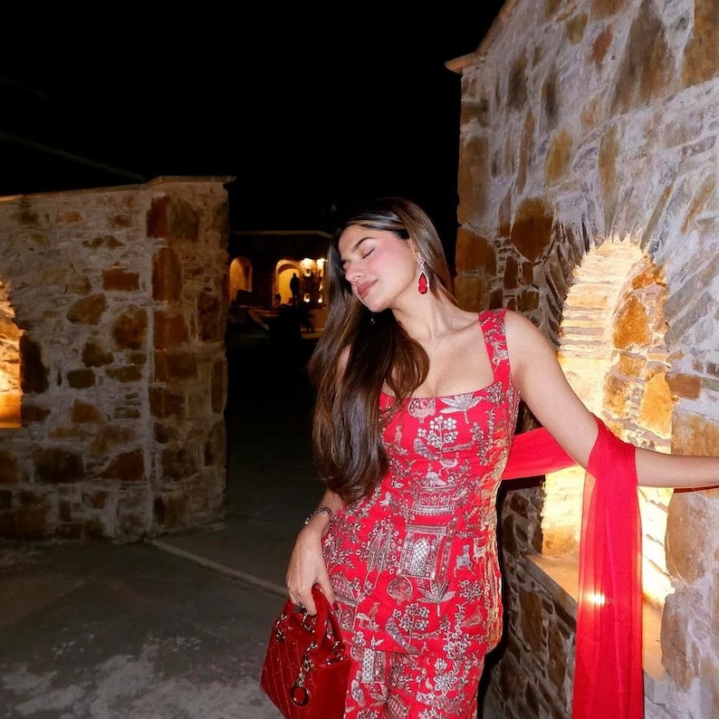 May include: A red ensemble featuring a patterned top and matching pants. The outfit includes a red handbag and a flowing scarf. The person is wearing red earrings. The background is a stone wall with archways.