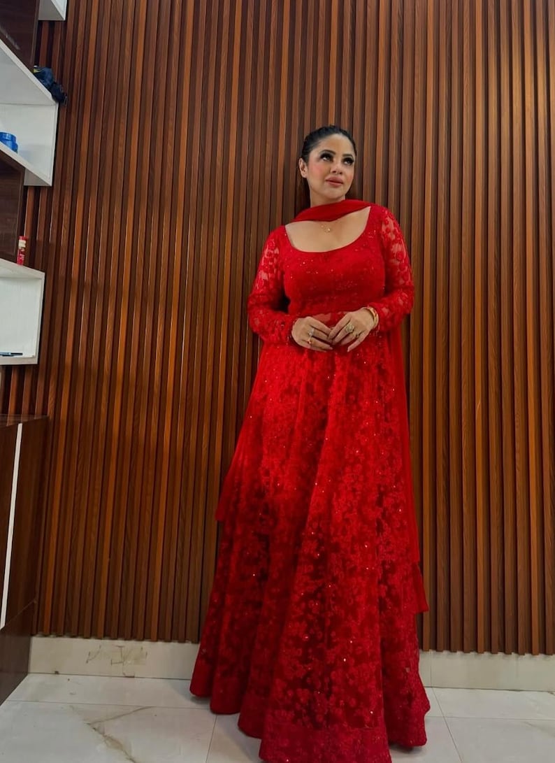 May include: A full-length red lace gown with long sleeves and a matching scarf. The dress features a square neckline and a tiered skirt. The background is a wooden wall.