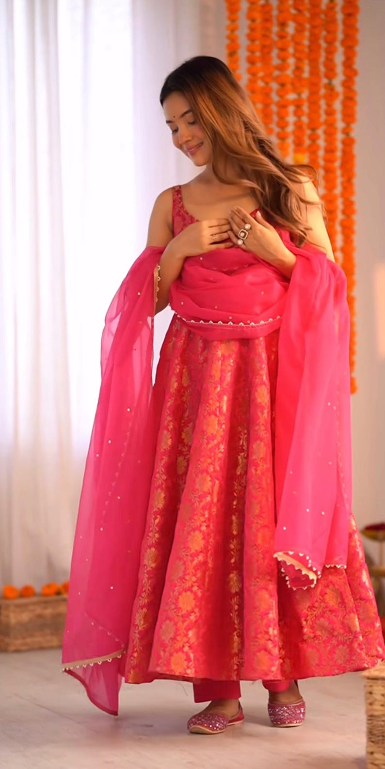 May include: A vibrant pink and gold floral patterned dress with a matching sheer scarf. The dress has a sleeveless top and a full skirt. The scarf is draped over the shoulders. The outfit is completed with matching pink shoes.