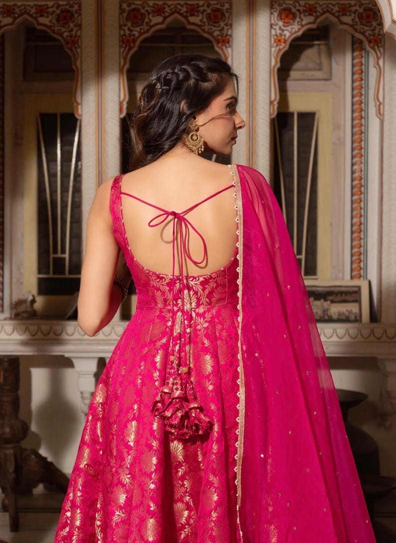 May include: A vibrant fuchsia-colored gown with gold floral patterns. The dress features a low back with tie detailing and a matching sheer scarf. The gown's design includes a tassel embellishment, creating an elegant and festive look.
