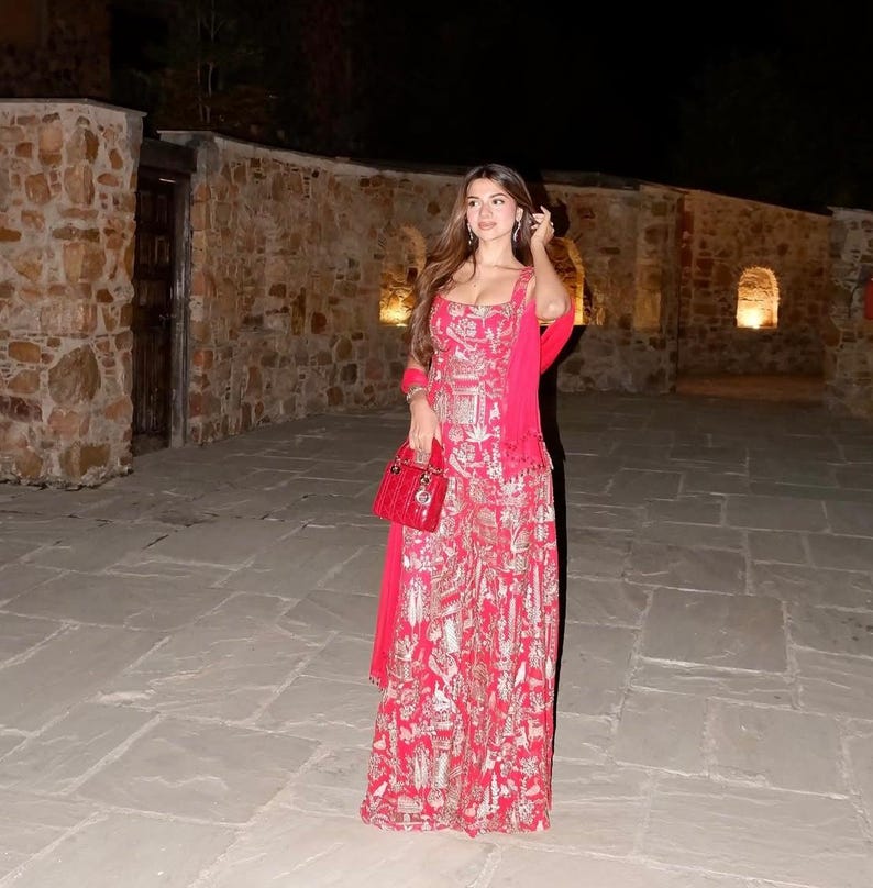 May include: A woman wearing a red and gold patterned dress with a matching scarf and a red handbag. The dress has a square neckline and spaghetti straps. The background features a stone wall and a paved walkway.