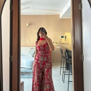 May include: A red jumpsuit with a matching scarf, featuring a detailed gold pattern. The jumpsuit has a square neckline and wide legs. The outfit is reflected in a mirror, showcasing the full length of the garment.