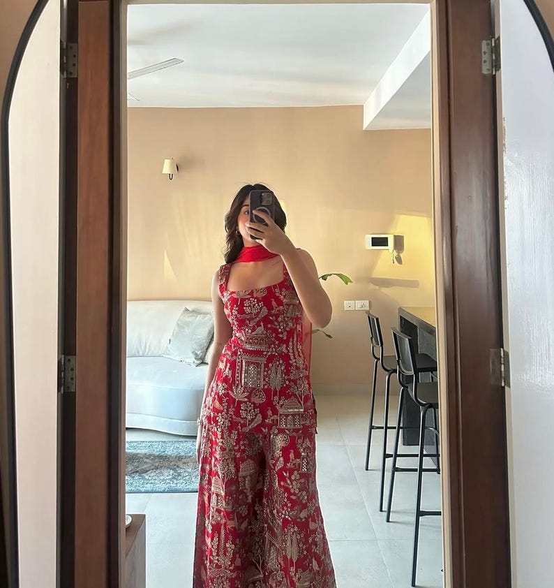 May include: A red jumpsuit with a matching scarf, featuring a detailed gold pattern. The jumpsuit has a square neckline and wide legs. The outfit is reflected in a mirror, showcasing the full length of the garment.