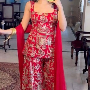 May include: A red embroidered outfit featuring a square-neck top and wide-leg pants. The outfit is adorned with gold embellishments and a matching red scarf. The ensemble is a traditional style, suitable for special occasions.