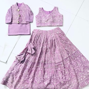 Puede incluir: Un conjunto de ropa para niños de color lila compuesto por una blusa sin mangas, una falda y una chaqueta. El atuendo está decorado con bordados y lentejuelas plateadas.